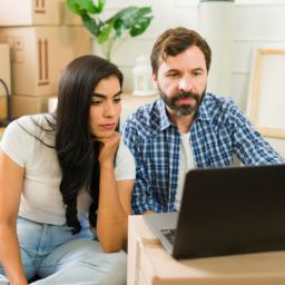 A worried couple sitting among moving boxes, looking at a laptop while reviewing their mortgage and foreclosure options