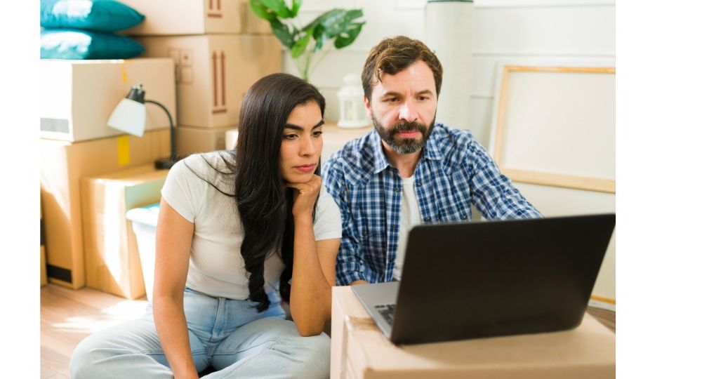 A worried couple sitting among moving boxes, looking at a laptop while reviewing their mortgage and foreclosure options