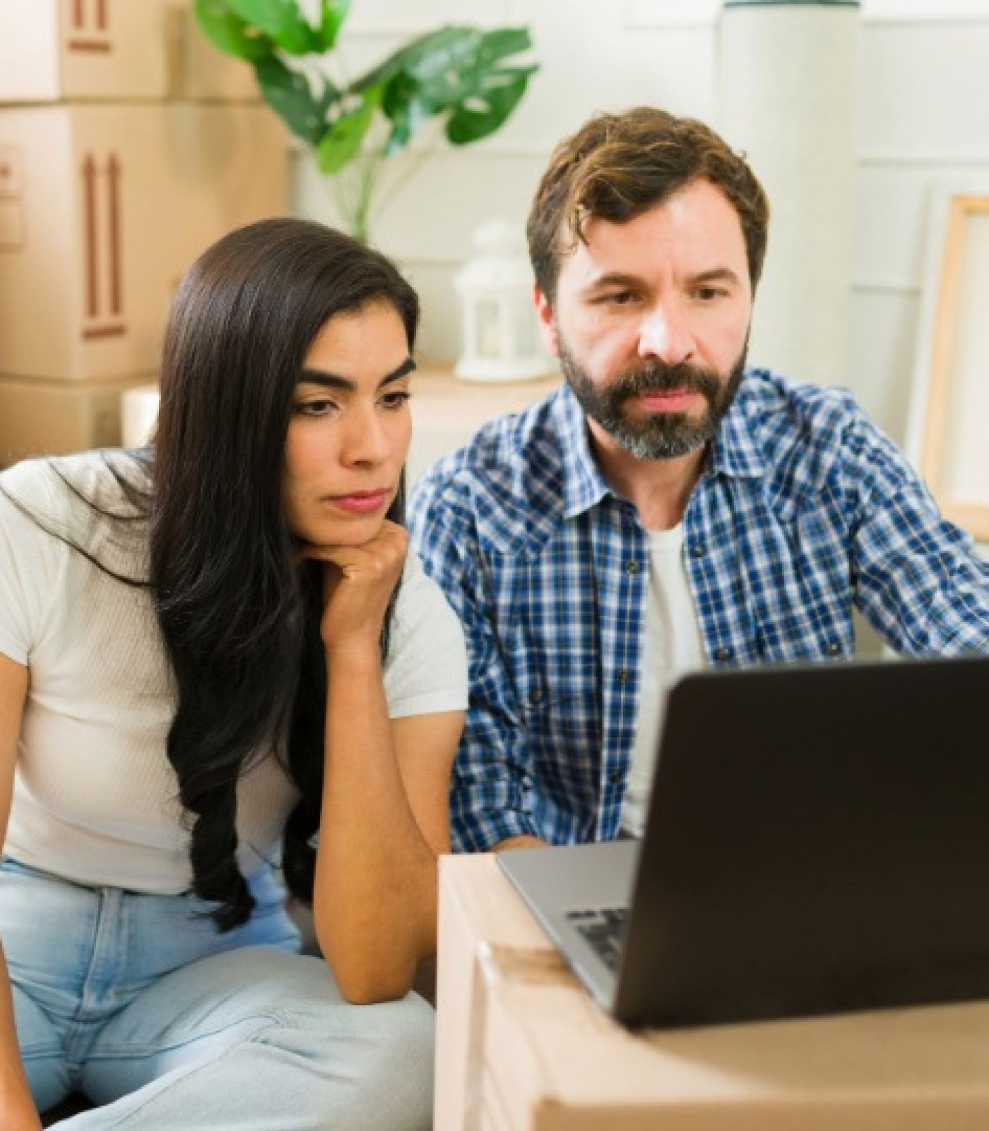 A worried couple sitting among moving boxes, looking at a laptop while reviewing their mortgage and foreclosure options