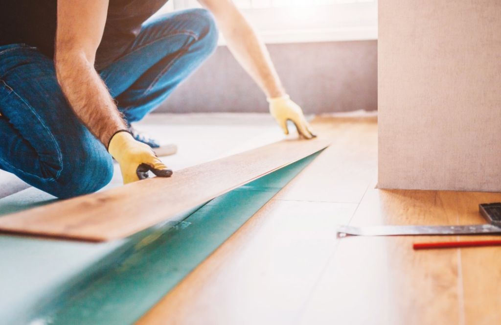 A person wearing work gloves kneels to fit a new plank of wood flooring.