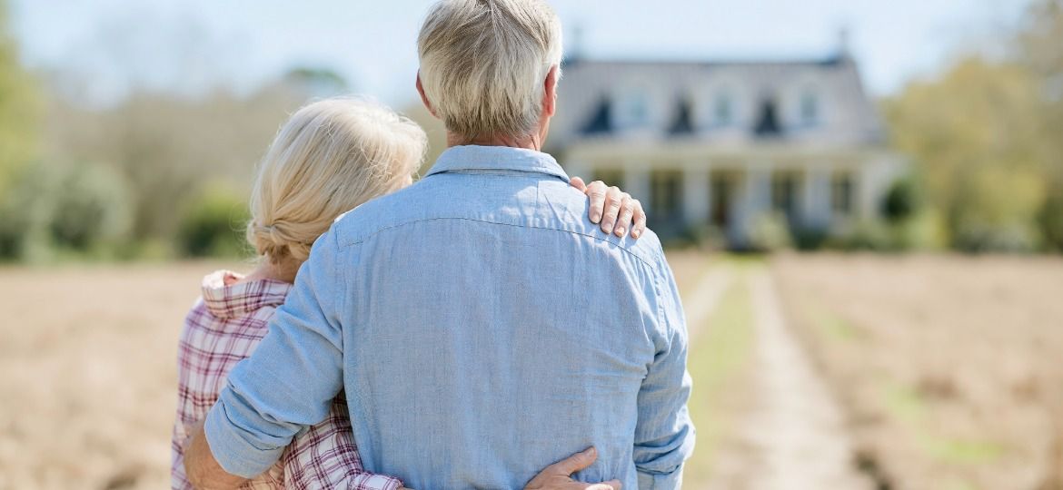 An older couple stand with their arms around each other, looking toward their large house.
