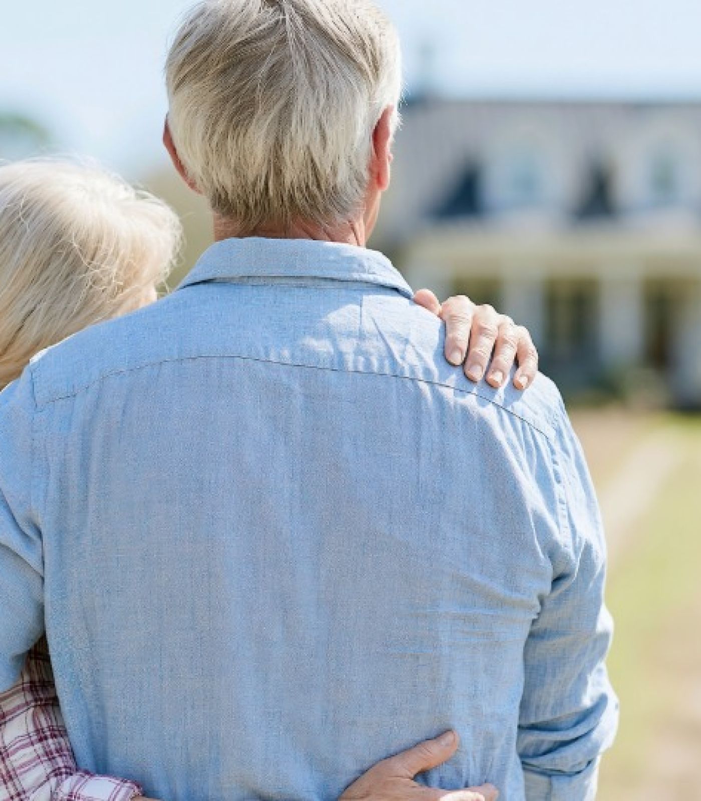 An older couple stand with their arms around each other, looking toward their large house.