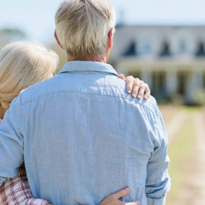An older couple stand with their arms around each other, looking toward their large house.